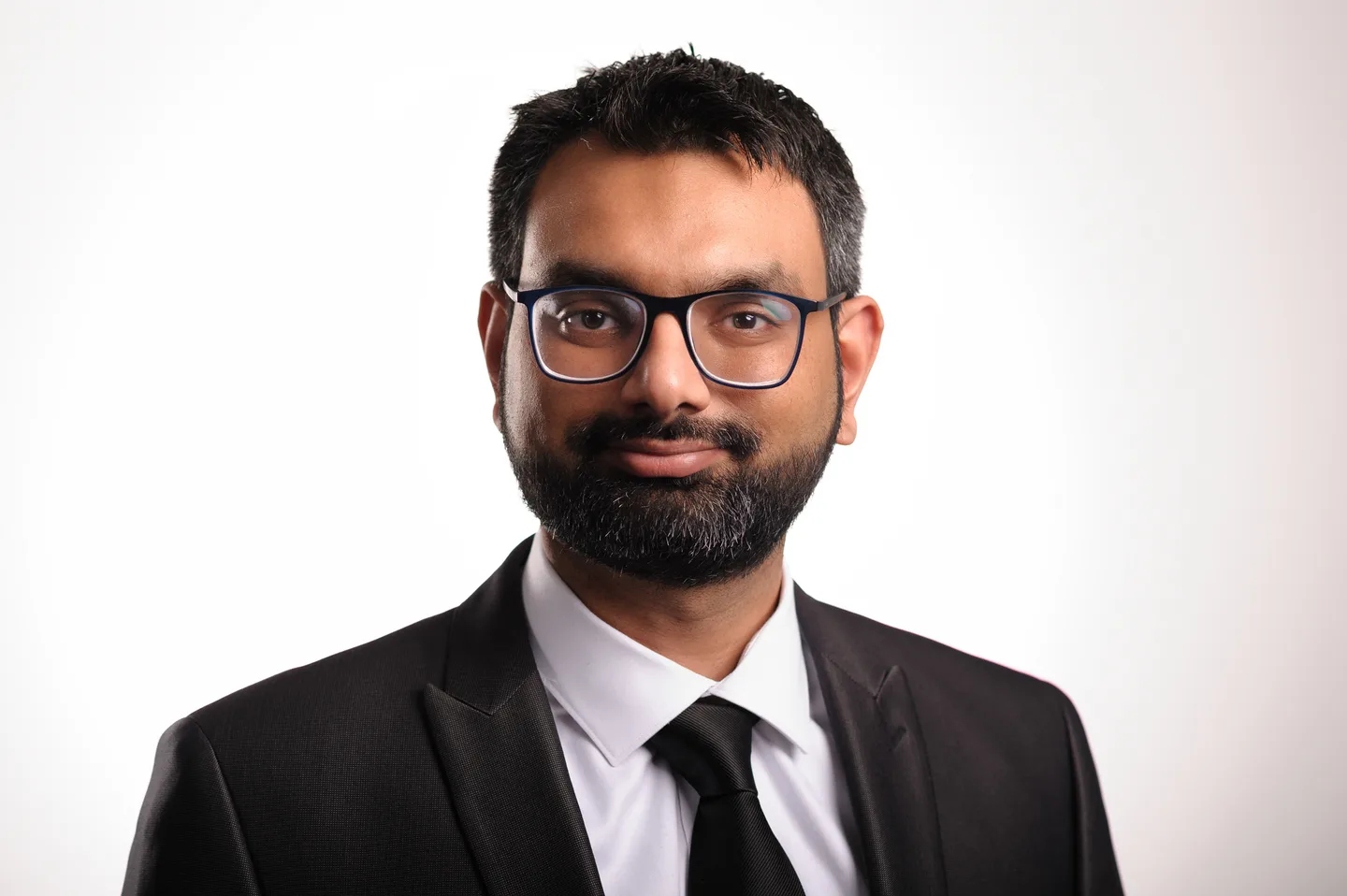 Studio headshot of a man with short dark hair, a full beard and glasses in a black suit and black tie