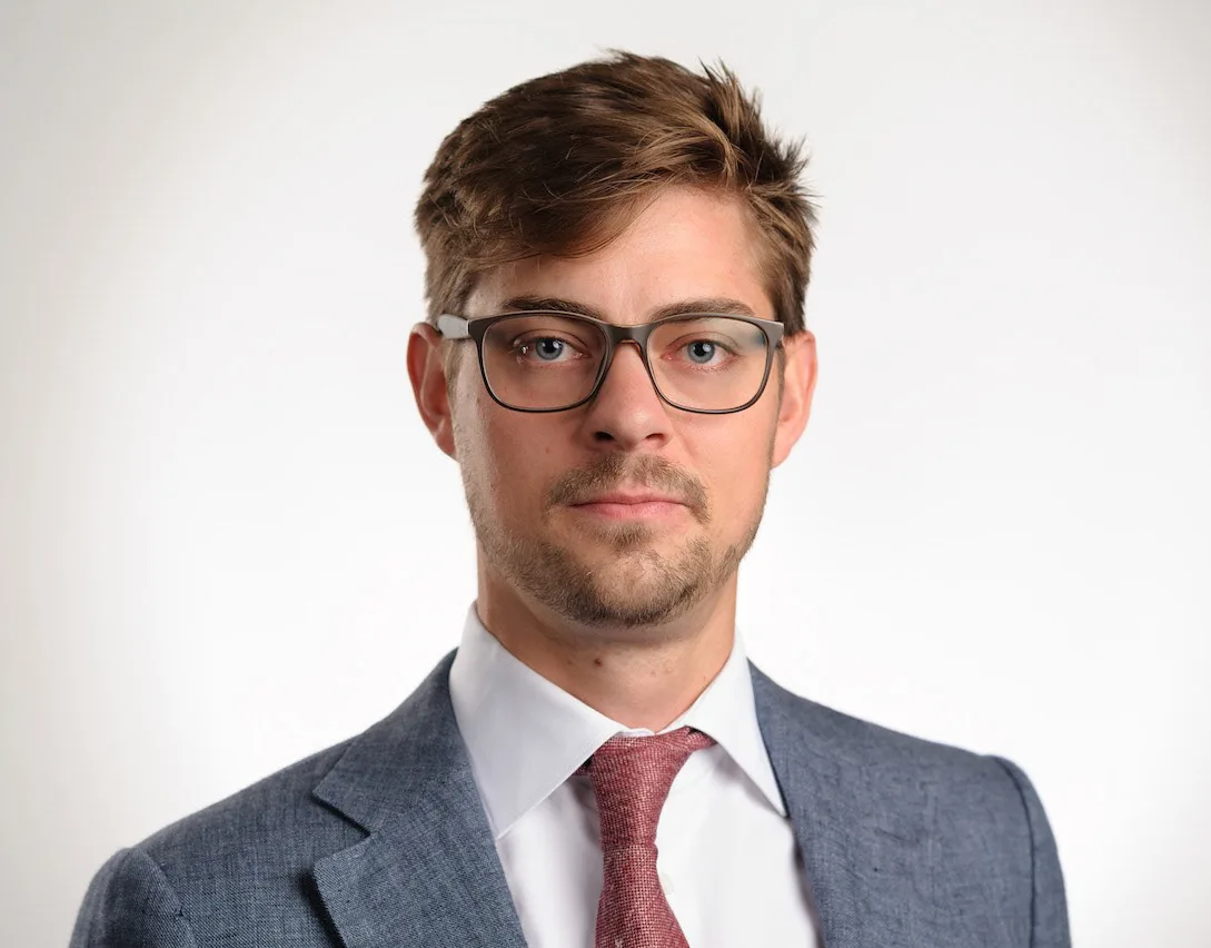 Studio headshot of a man with tousled brown hair and glasses in a grey-blue jacket and red tie
